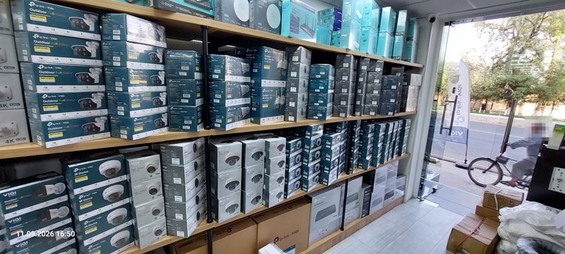 Стеллаж с камерами TP-Link VIGI (bullet, IR Dome, turret), внизу коробка VIGI 16 Channel PoE+ NVR, сверху Wi-Fi-усилители; справа через стекло двери виден велосипедист на улице