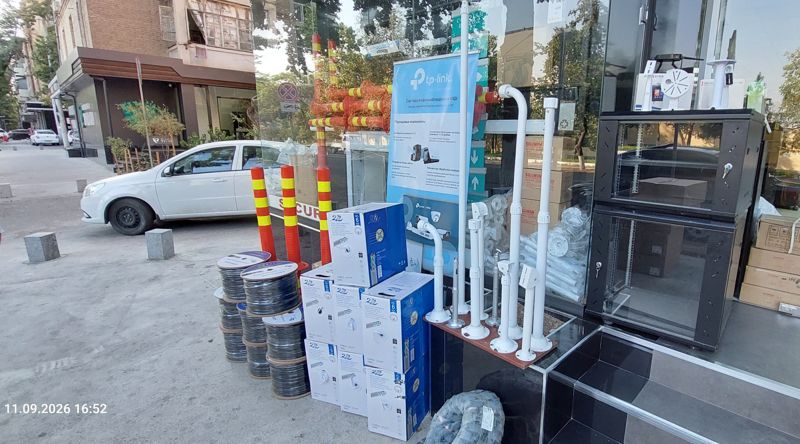Витрина у входа на улице: катушки уличного кабеля CAT6 UTP, коробки кабеля CAT6, белые стойки и кронштейны для камер, чёрные настенные телекоммуникационные шкафы, плакат TP-Link VIGI, оранжевые гибкие столбики