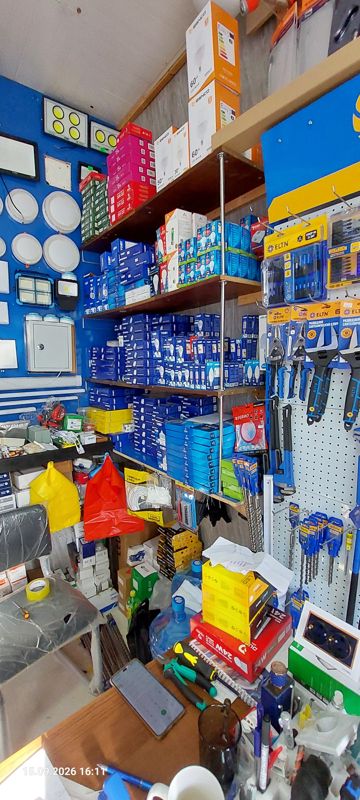 Электротовары: стеллаж со светодиодными лампами в коробках (Tobi, Esnco 50–60 W LED Bulb и др.), на синей стене — светодиодные прожекторы, встраиваемые панели-светильники, электрощиток; справа стенд ELTN с инструментом