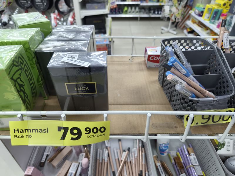 Верхняя полка стеллажа: парфюмерия Avon Luck и другие, групповая табличка «Hammasi / Всё по 79 900 so'mdan»