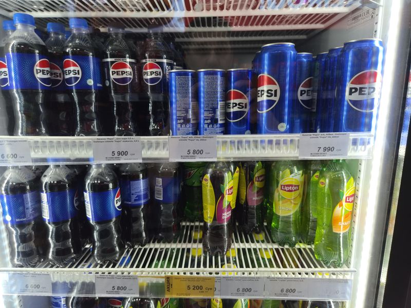 Полка холодильника с Pepsi в бутылках и банках, Lipton и Mountain Dew; белые ценники и один жёлтый акционный