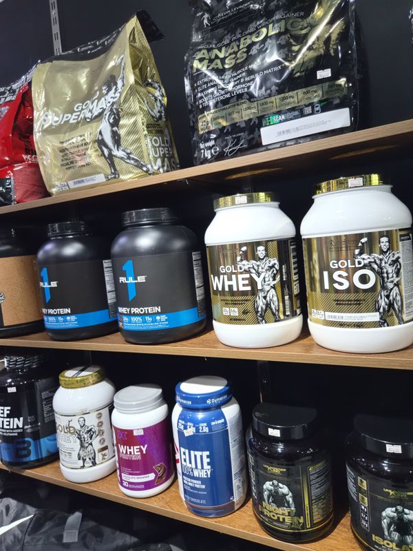 Стеллаж с гейнерами и протеинами: Gold Super Mass, Anabolic Mass, Rule One, Gold Whey, Elite Whey