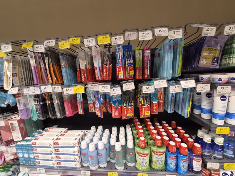 Зубные щётки Colgate, Splat, Biomed и Bonna на крючках в два ряда, ополаскиватели и зубные пасты Splat и R.O.C.S. внизу; белые и жёлтые ценники