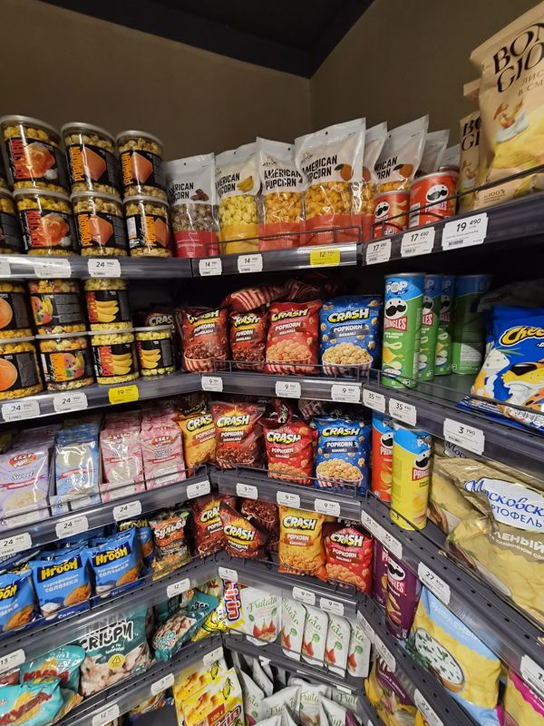 Угловой стеллаж снеков: попкорн Real American, Bisyor, Crash, Eazy Pop; чипсы Pringles, Bon Giorno, Cheetos; белые и жёлтые ценники