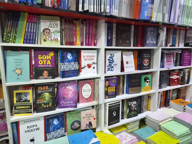Азбука: стеллаж с книгами на узбекском и русском языках, сверху папки-файлы A4, внизу стопки школьных тетрадей