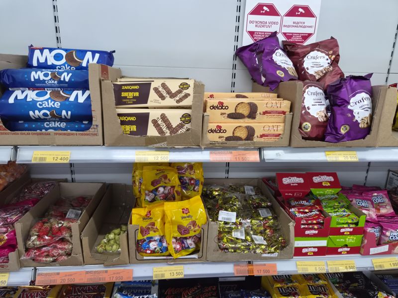 Havas: полка с печеньем Moon cake, Zarqand Shedevr, Delato Oat и Crafers, ниже конфеты в картонных лотках, Skittles и Mexxi Corn; вверху наклейки о видеонаблюдении