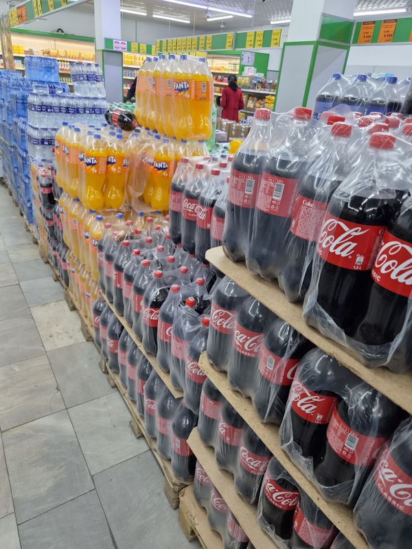 Паллеты с газированными напитками и водой в торговом зале Havas: Coca-Cola Classic 1,5 л, Fanta, вода в упаковках; на подвесных планках над проходом жёлтые и оранжевые ценники