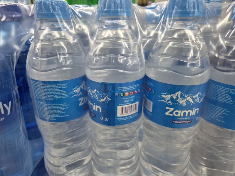 Упаковки питьевой воды Zamin негазированной 0,5 л в плёнке, на задней этикетке штрихкод; слева край упаковки Family
