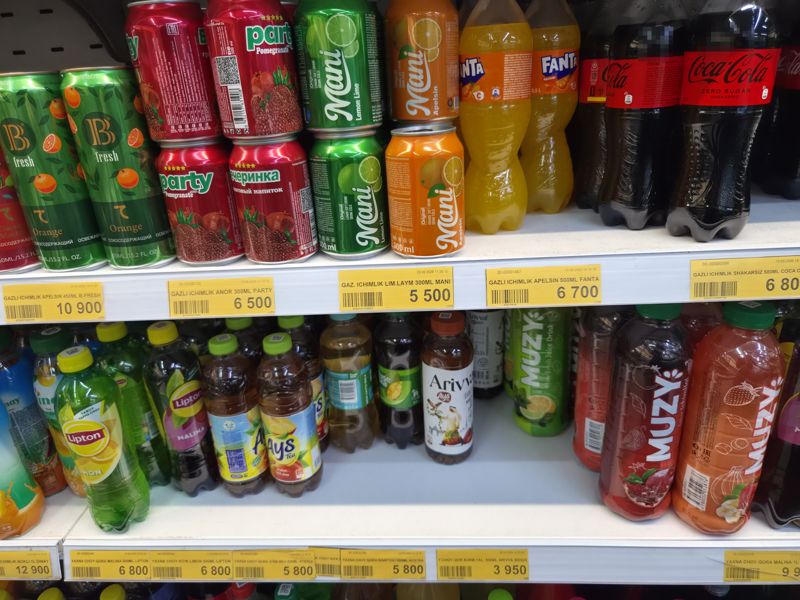 Две полки: сверху банки B-Fresh, Party, Mani и бутылки Fanta, Coca-Cola Zero; ниже бутылки холодного чая Lipton, Aystea, Arivva и соков Muzy; под полками жёлтые ценники