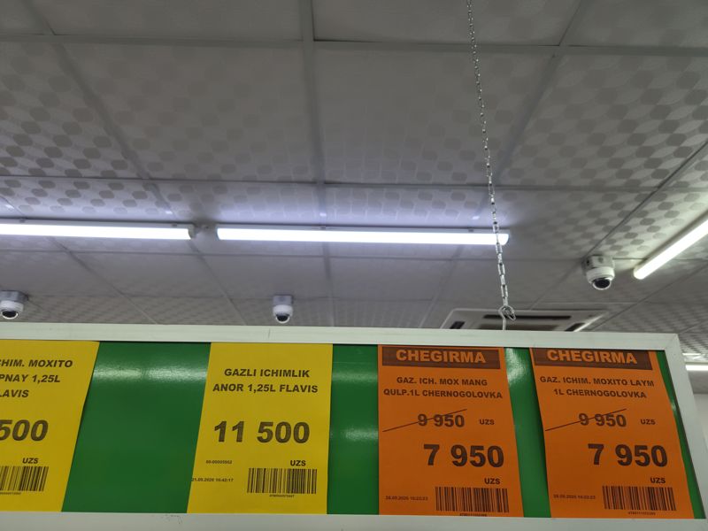 Ценники под потолком над стеллажами: жёлтый ценник Flavis (гранатовая газировка 1,25 л) и два оранжевых ценника Chegirma на Chernogolovka 1 л (мохито), слева обрезан ещё один жёлтый, вверху купольные камеры видеонаблюдения