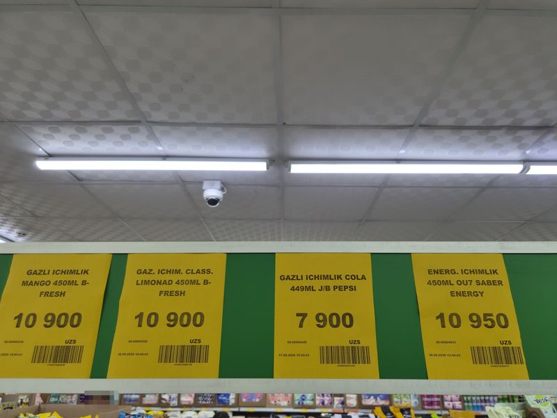 Жёлтые ценники под потолком на газировку и энергетик в банках по 450 мл: B-Fresh манго, B-Fresh классический лимонад, Pepsi Cola, Saber Energy; вверху камера видеонаблюдения, внизу товары в коробках и на полках