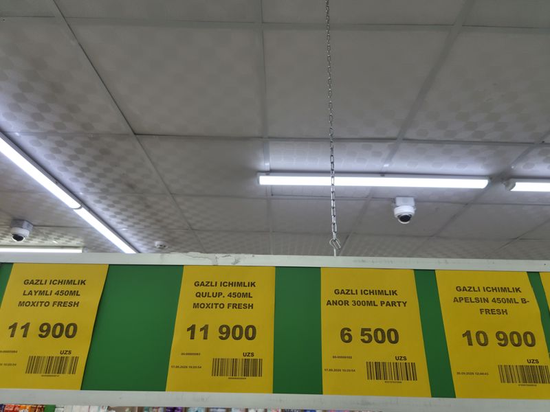 Жёлтые ценники под потолком на газировку: Moxito Fresh лайм и клубника 450 мл, Party гранат 300 мл, B-Fresh апельсин 450 мл; внизу видны полки с бытовыми товарами и банки Coca-Cola, вверху камеры видеонаблюдения