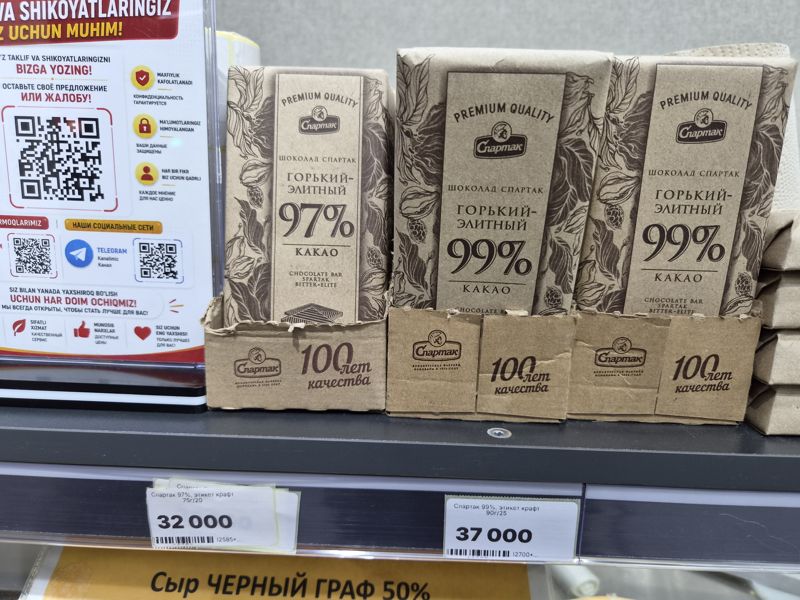 Шоколад Спартак горький элитный 97% и 99% какао в крафтовой упаковке на полке, слева стойка с QR-кодами обратной связи и соцсетей магазина, внизу край жёлтого плаката «Сыр Чёрный граф 50%»