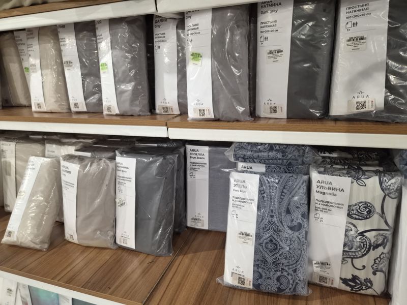 Полки с постельным бельём ARUA: простыни на резинке «Далия» Light beige и Grey, «Натмина» Dark grey, комплекты «пододеяльник и наволочки» «Далия», «Натмина» Dark grey, «Копелла» Blue Jeans, «Этель» Dark blue, «Ульвина» Magnolia; на упаковках наклейки с ценой, часть выделена зелёным маркером