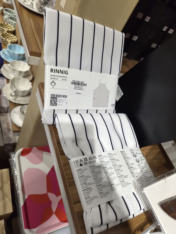 Фартук IKEA RINNIG в белую и тёмно-синюю полоску, 69×85 см, с наклейкой цены; на заднем плане чашки, тарелки и поднос