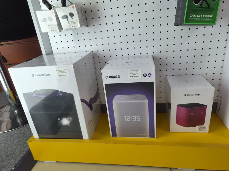 Три коробки умных колонок Яндекс на жёлтой подставке — Станция Макс с Zigbee, Станция 3 с Zigbee, Станция Миди с Zigbee; вверху проводные наушники Xiaomi Mi In-Ear и автомобильное зарядное Infinix