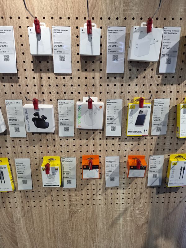 Стенд аксессуаров на перфорированной панели в офисе Ucell: адаптеры питания Apple, наушники Redmi Buds 4 Pro и Buds 4, Baseus Bass BP1 Pro, колонки Xiaomi, кабель Baseus с ценниками-карточками