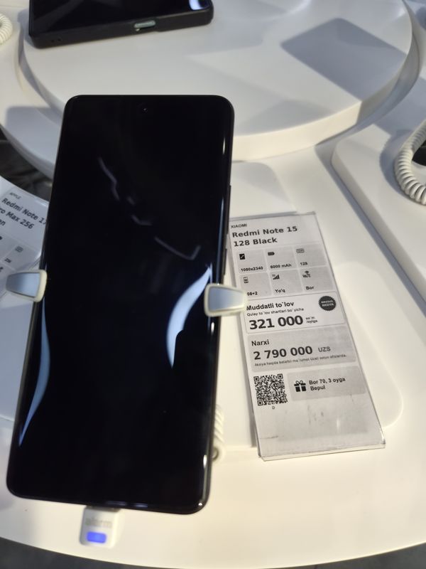 Демонстрационный смартфон Redmi Note 15 на белой стойке Ucell с ценником «XIAOMI Redmi Note 15 128 Black»