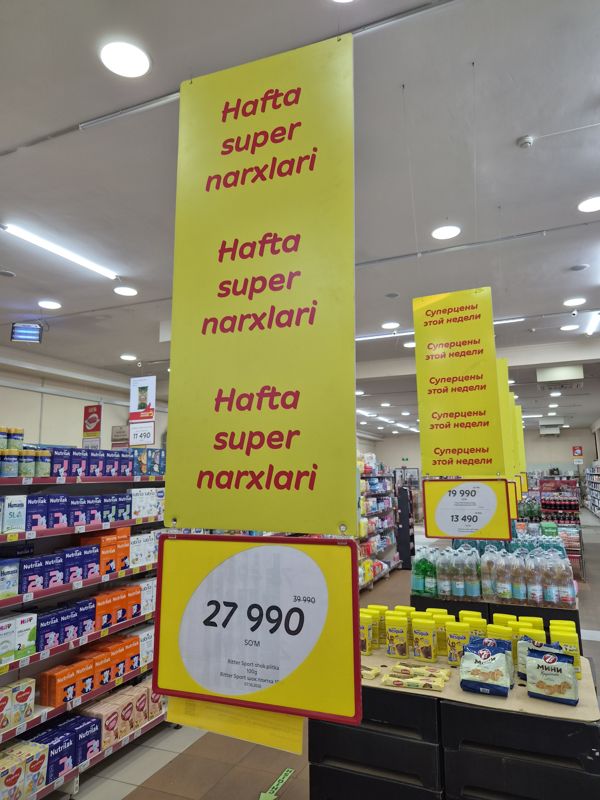 Подвесной постер «Hafta super narxlari» над горкой: ценник на шоколад Ritter Sport, вдали ценник на круассаны 7 Days и вафли Яшкино