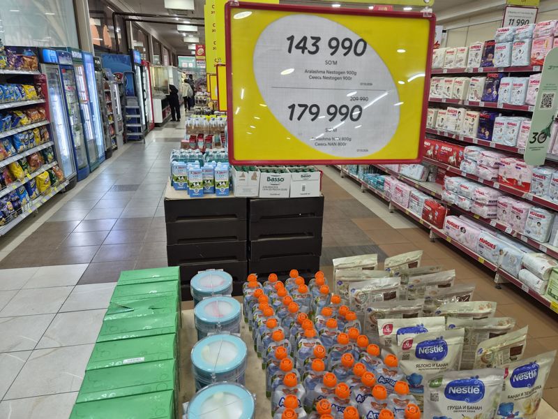 Стол с детским питанием под подвесным ценником: детские смеси Nestogen 900 г и NAN 800 г; на столе детские каши Nestle, пюре Агуша, смеси NAN и Nestogen; сзади поддон с молоком Милка и оливковым маслом Basso