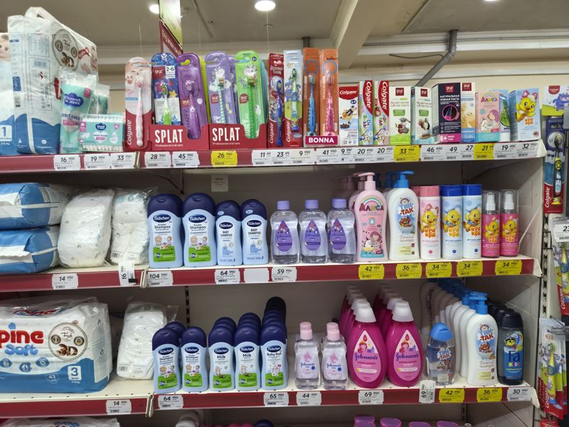 Стеллаж детской гигиены: сверху детские зубные щётки и пасты (Splat, Colgate, Bonna, Алиса, Кря-Кря), ниже детские шампуни и масла (Bubchen, Johnson's, Тик-Так, Алиса, Кря-Кря), подгузники слева