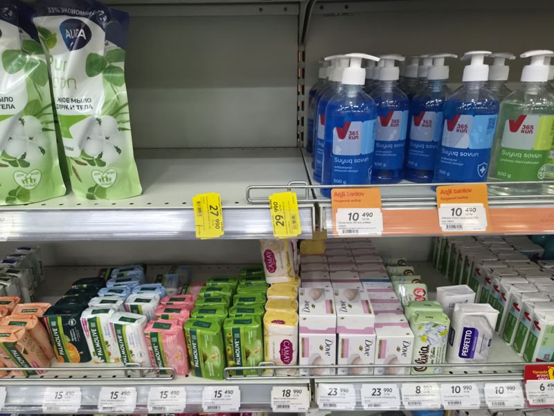 Жидкое мыло: пакеты Aura и бутылки 365 kun (антибактериальное), ниже мыло Palmolive, Camay, Dove, Love Nature и Perfetto; на полке оранжевая плашка «Aqlli tanlov» и жёлтые ценники, повёрнутые вертикально