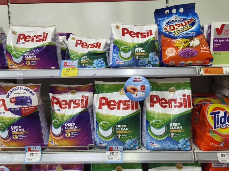 Полки со стиральными порошками Persil Deep Clean Plus (Color, Свежесть от Vernel), Kopum Plus и Tide; ценники с наклейками +1 o'yinchoq, на упаковке круглые стикеры «7% keshbek» и Persil