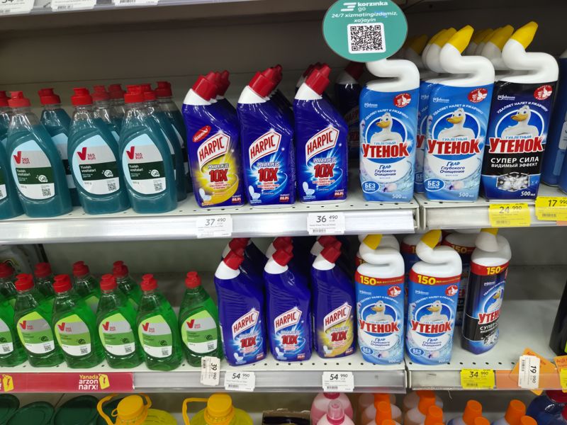 Полки со средствами для унитаза Harpic Power Plus и «Утёнок», внизу слева гели для посуды 365 kun; над полкой подвесной круг Korzinka Go с QR