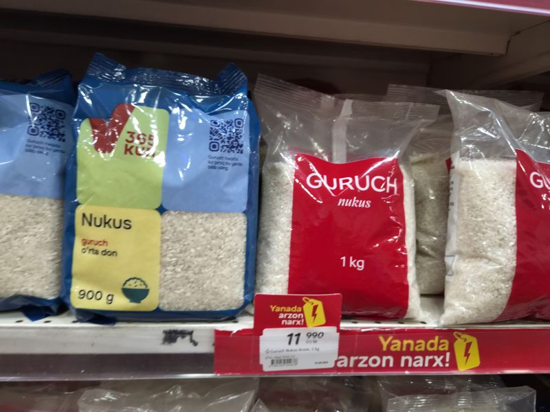 Полка с рисом: пачки 365 kun Nukus 900 г (синие) и Guruch Nukus 1 кг (красные); под красными пачками красная планка «Yanada arzon narx!» и белый ценник