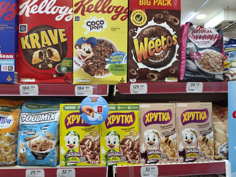 Стеллаж с готовыми завтраками: Kellogg's Krave, Coco Pops, Weetos Big Pack, хлопья «Гармония» AXA, Goodmix «Чуррос Вайб», Хрутка (подушечки с бананом и шоколадные); белые полочные ценники, подвесной круг «Покупайте Хрутка» с игрушкой