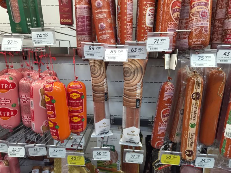 Колбасный отдел: ценники на Rozmetov, Delmark, Sherin; внизу ценники на вяленое мясо Del Jerky и нарезки Toxtaniyoz Ota; справа жёлтые ценники «Hafta super narxlari»
