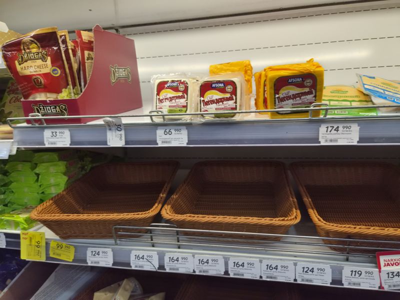 Сырная витрина: Dziugas Hard Cheese и сыры Afsona «Легендарный» на верхней полке, пустые плетёные корзины над рядом ценников весовых сыров Mlekovita, Беловежские; слева сверху видны пачки сыра, справа красная плашка «Narxiga javob!»