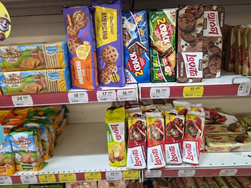 Два стеллажа с печеньем: Коровка, Zarqand Americano, Roshen 2 Crack и Lovita (Dark Cocoa, Jelly Cookies, Blondie Brownie), Яшкино Jaffa Cake; ценники на красных кромках, жёлтые акционные «Hafta super narxlari»