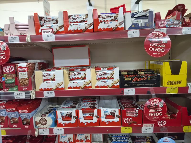 Полки с шоколадными батончиками: Kinder Bueno, Kinder Delice, KitKat Chunky, Mars, Nuts, Otto, Nestle, плашки «Narxiga javob!» и круглые наклейки KitKat F1
