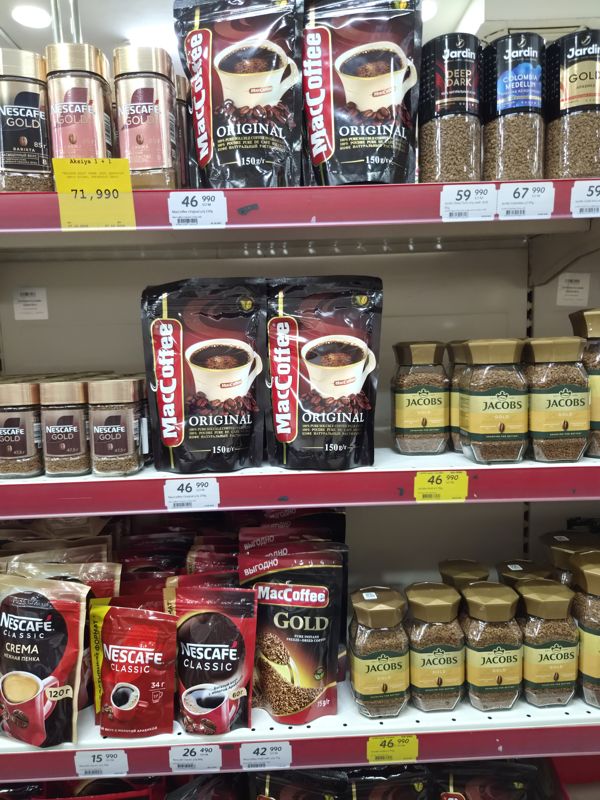 Полки с растворимым кофе: MacCoffee Original в пакетах, Jacobs Gold в банках, Jardin, Nescafe Gold, Nescafe Classic и MacCoffee Gold; карточка «Aksiya 1 + 1» и жёлтые ценники «Hafta super narxlari»