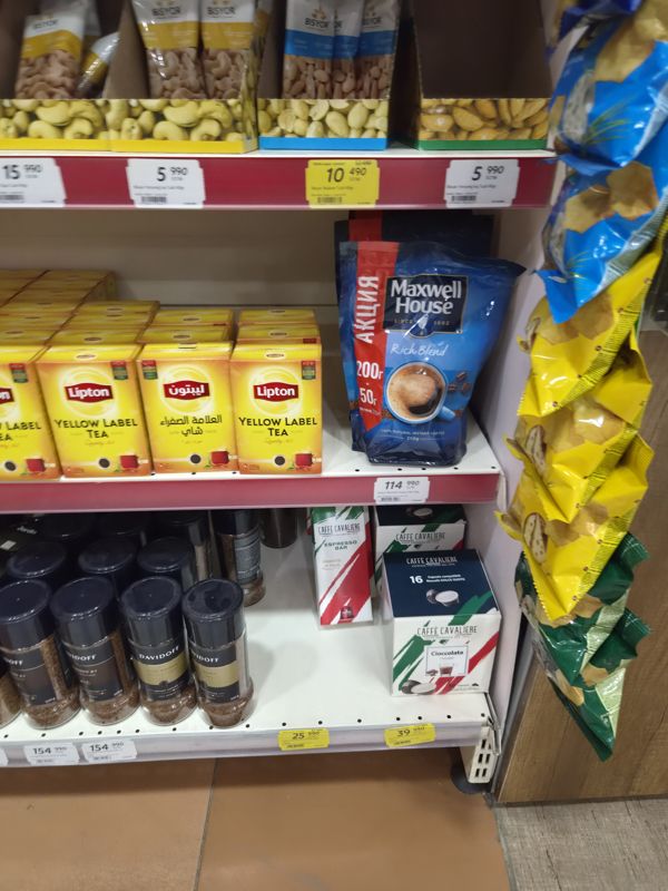 Нижние полки стеллажа: орешки Bisyor в коробках, чай Lipton Yellow Label, Maxwell House Rich Blend в пакете, банки Davidoff и капсулы Caffe Cavaliere; белые и жёлтые ценники