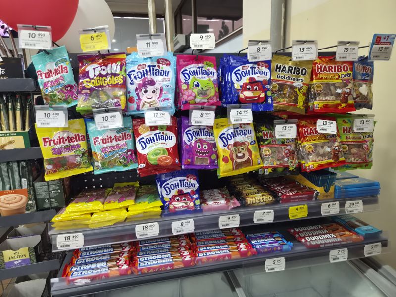 Стойка с жевательным мармеладом Haribo, Бон Пари, Fruittella, Trolli и Chupa Chups на крючках; ниже полки с батончиками и шоколадом Strobar, KitKat, Metro, AXA, Goodmix; кадр с движением