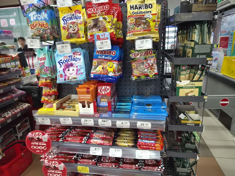 Торец стеллажа с мармеладом Haribo, Бон Пари, Fruittella и Trolli на крючках, ниже шоколад и батончики KitKat, Nestle, Metro, Strobar; плакат KitKat F1 «Yutib oling»; слева кассиры