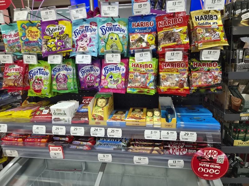 Стойка с мармеладом Haribo, Бон Пари, Fruittella и Chupa Chups на крючках; ниже плитки и батончики Алёнка, Ritter Sport, Damak, Рахат, Snickers, Bounty, Twix Xtra, KitKat Chunky; красная плашка «Narxiga javob!»