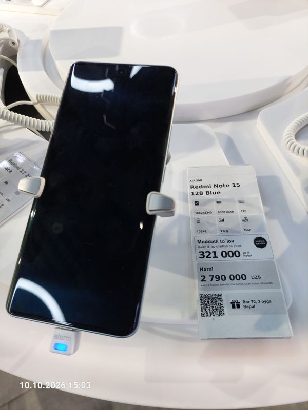 Смартфон Xiaomi голубого цвета на витрине Ucell в держателе с сигнализацией, рядом ценник-карточка: XIAOMI Redmi Note 15 128 Blue, 2 790 000 сум