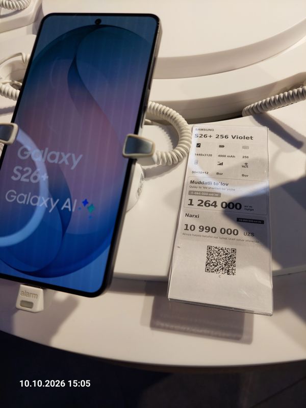 Смартфон Samsung Galaxy S26+ в держателе с сигнализацией на витрине Ucell, рядом ценник-карточка: SAMSUNG S26+ 256 Violet, 10 990 000 сум, перечёркнутая прежняя цена 12 990 000
