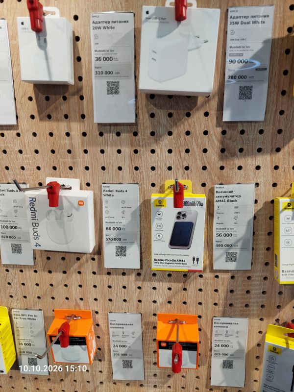 Стенд аксессуаров на перфорированной панели в офисе Ucell: адаптеры питания Apple 20W и 35W Dual, наушники Redmi Buds 4, внешний аккумулятор Baseus PicoGo AM41, колонки Xiaomi и Baseus Bass BP1 Pro с ценниками-карточками
