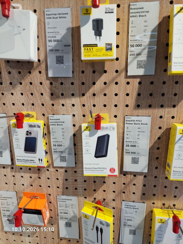 Стенд аксессуаров на перфорированной панели в офисе Ucell: адаптер питания Apple 35W Dual, внешние аккумуляторы Baseus AM41 и EnerFill FP21, колонка Xiaomi, кабель Baseus с ценниками-карточками