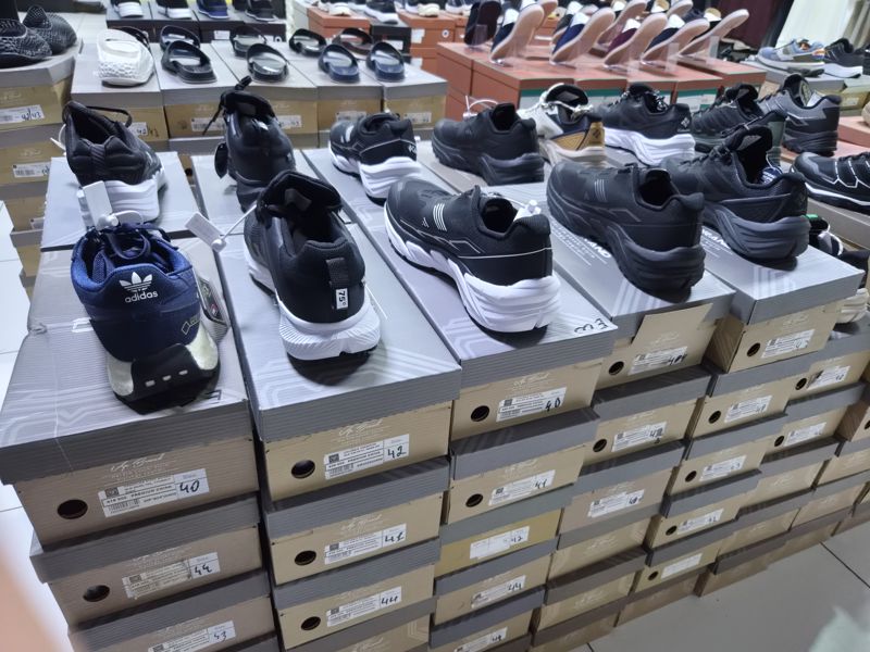 Стойка кроссовок на серых коробках: слева тёмно-синие adidas с GORE-TEX, правее чёрные и чёрно-белые Columbia; на торцах коробок наклейки с моделью, ценой и размером, вверху витрина с сандалиями и другой обувью