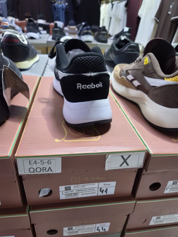 Кроссовки Reebok на розовато-коричневой коробке Per La Umma в стопке; на торце наклейка с моделью, ценой и размером, на соседней коробке рукописная этикетка «E4-5-6 QORA»; справа коричневые кроссовки, на фоне полки и вешалки с одеждой