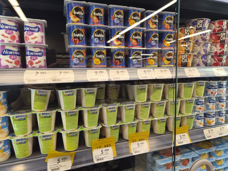Холодильная витрина с йогуртами: «Нежный» 5%, Fruttis 5%, Epica, Campina «Легкий» (в зелёных стаканах Ehrmann «Нежный»), справа «Нежный» 1,2% и Musaffo; ценники на кромках, жёлтые плашки «Aksiya 2 + 1» под ценниками Campina