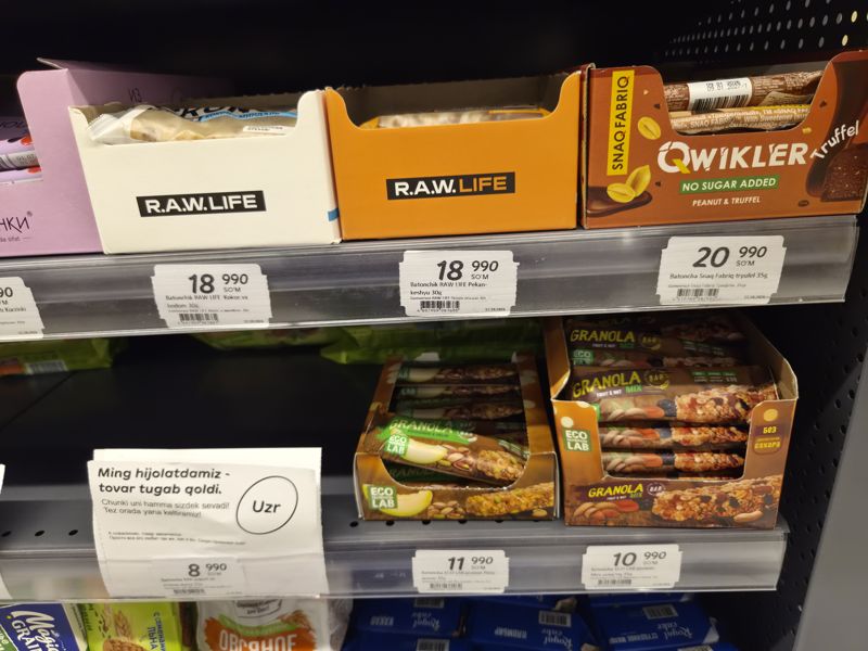 Полки с батончиками: R.A.W. Life, Snaq Fabriq Qwikler, ECO LAB Granola и AXA; на нижней полке карточка «Tovar tugab qoldi»