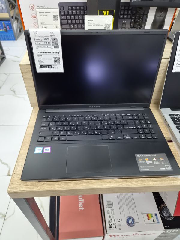 Ноутбук ASUS Vivobook Go 15 на деревянной подставке, на крышке два ценника; выше — ценники 2E на комплект клавиатуры и игровой коврик, снизу коробки Moulinex и Braun