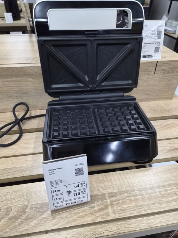 Сэндвичница Braun SM5005 (гриль/вафельница) с открытой крышкой на деревянной полке, перед ней белый ценник; справа в глубине ценник тостера Tefal