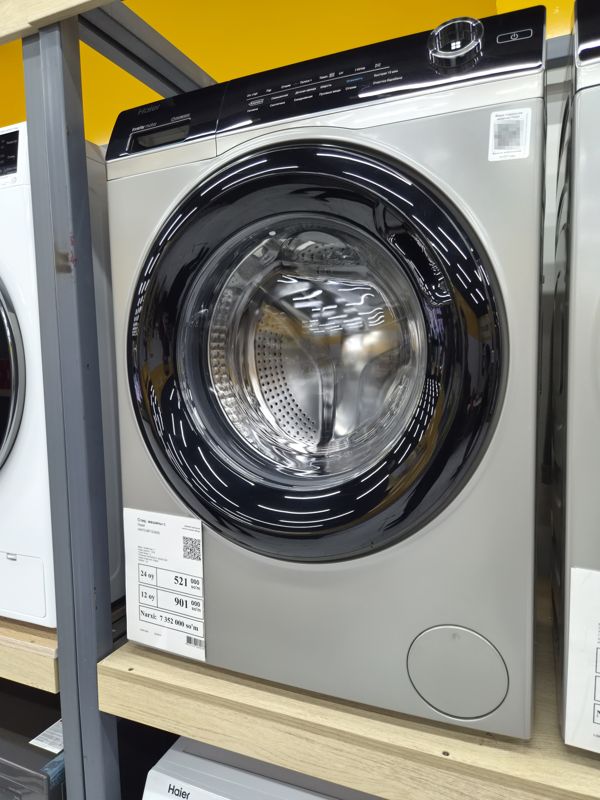 Стиральная машина Haier с фронтальной загрузкой серого цвета на полке магазина электроники, ценник на кронштейне